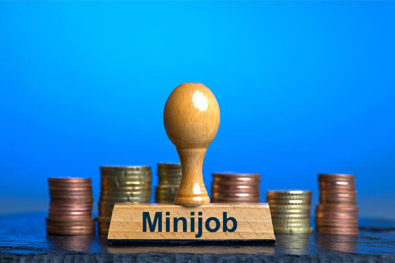 Ein Holzstempel mit der Aufschrift 'Minijob' vor mehreren Stapeln mit Euro / Cent-Münzen 