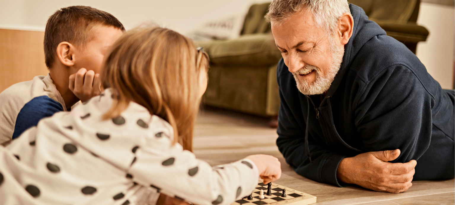 Grandfather looking at girl playing chess lying on floor at home