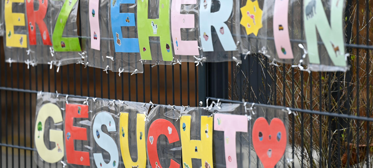 Mit bunten Buchstaben sind die Worte „Erzieher*in gesucht“ geformt, die an einem Zaun einer Kindertagesstätte befestigt sind