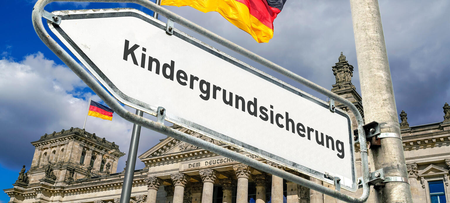 Ein weisses Hinweisschild an einem Laternenmast mit abgeblaetterter Farbe am Rand und der Aufschrift 'Kindergrundsicherung' als Wegweiser nach links vor dem Berliner Reichstagsgebaeude mit wehenden Deutschlandflaggen