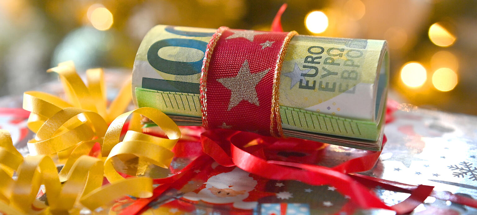 Symbolfoto Weihnachtsgeld: Zusamengerollte Euroscheine vor Weihnachtsbaum mit Schleife.