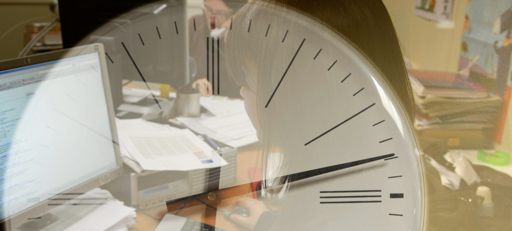 Die Abbildung zeigt eine große Uhr, im Hintergrund Bürobeschäftigte.